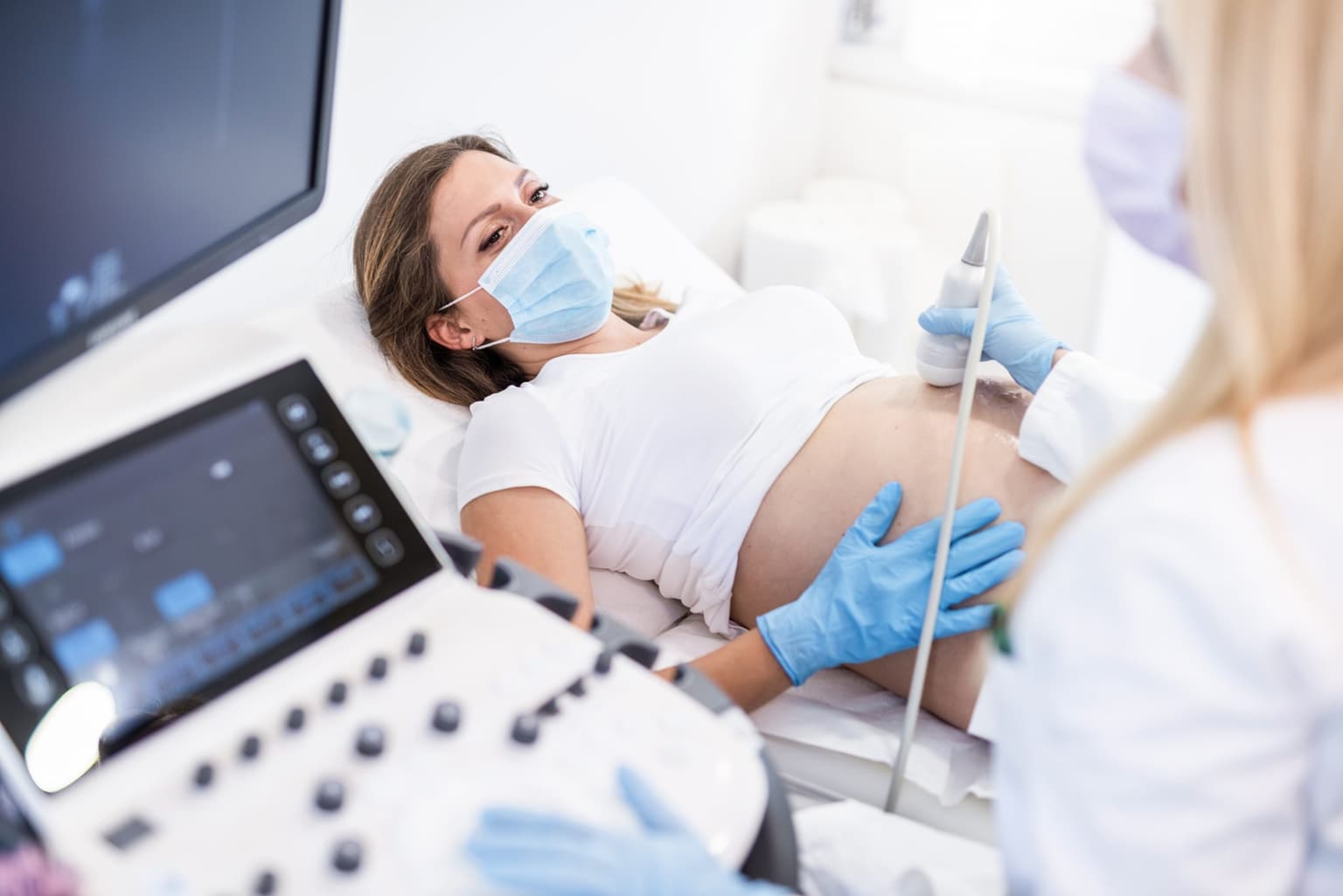 pictured is a woman having an ultrasound scan on her baby bump, she's wearing a mask and a white t-shirt and there's an ultrasound technician present